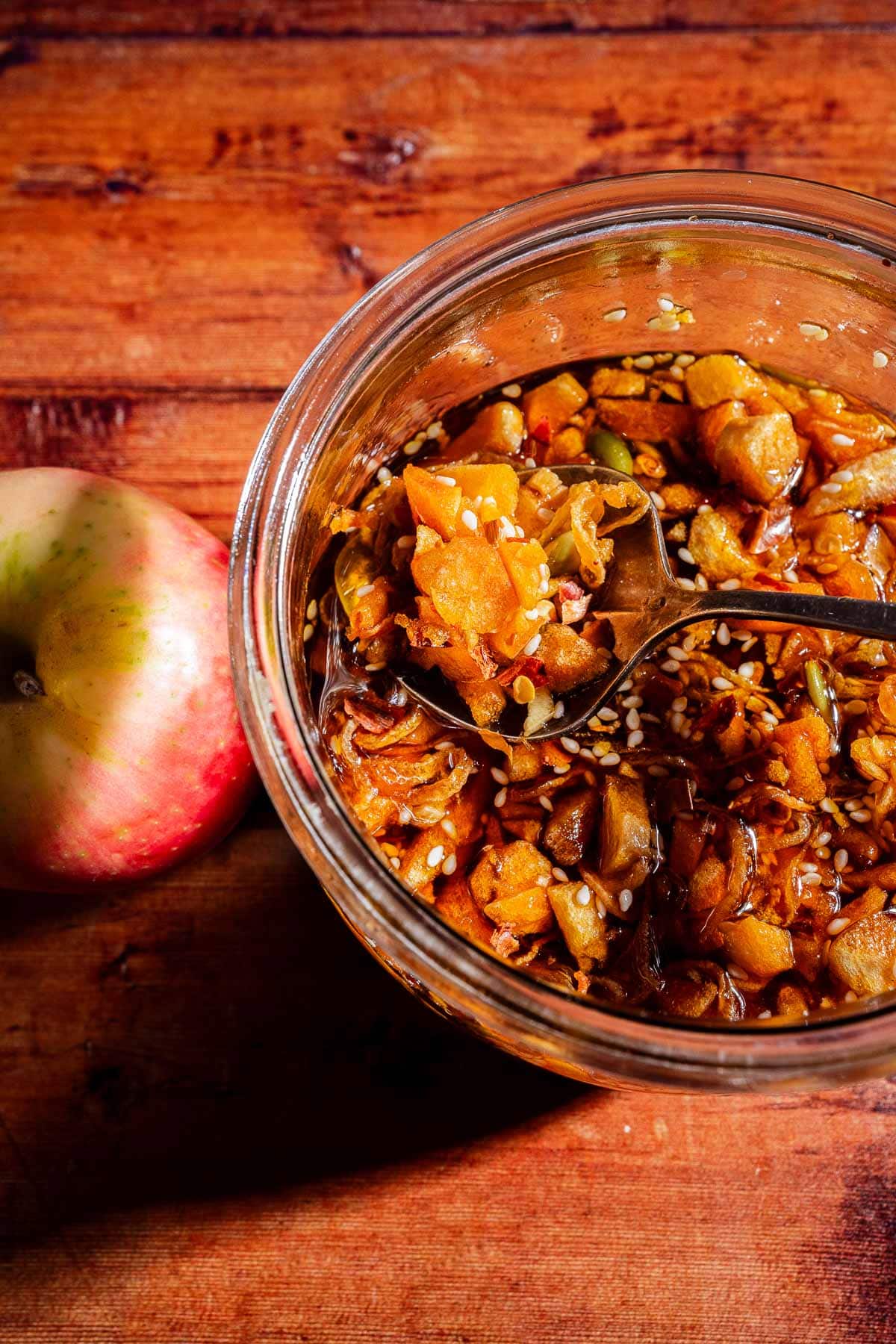 A glass bowl of chunky apple chutney with seeds, infused with a hint of apple chili crisp, sits on a wooden surface next to a whole apple. A spoon rests inside, ready for serving.