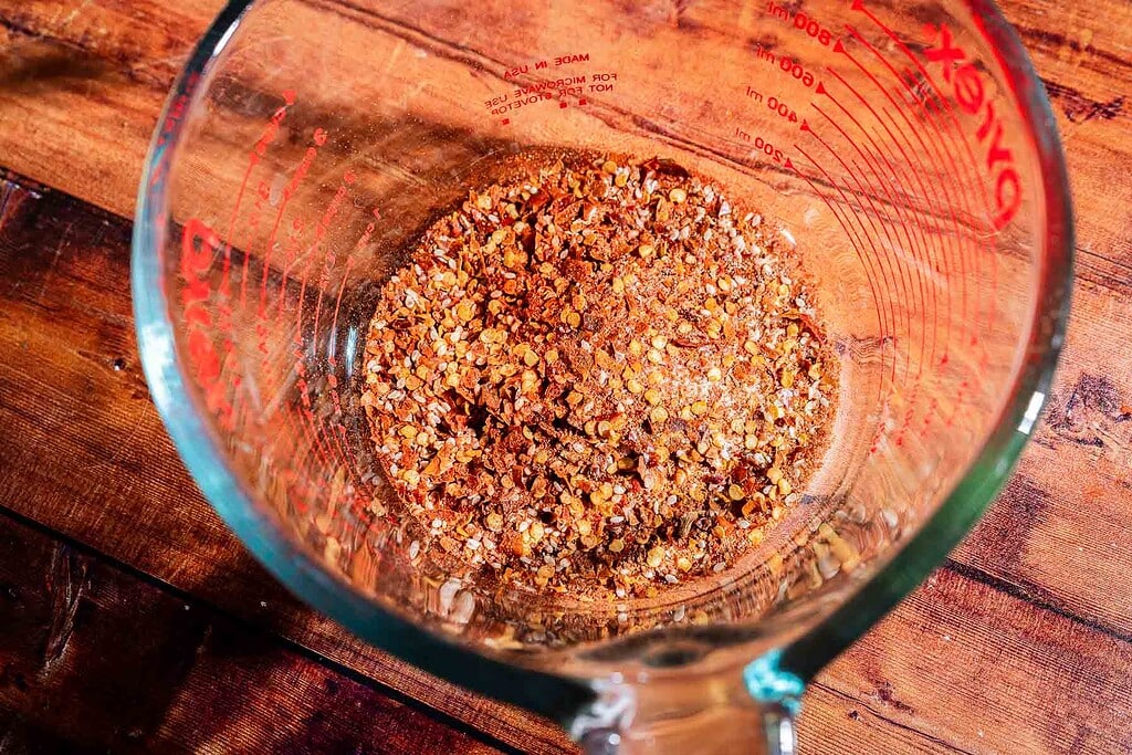 A glass measuring cup filled with a dry spice blend, inspired by the flavors of apple chili crisp, sits on a wooden surface.