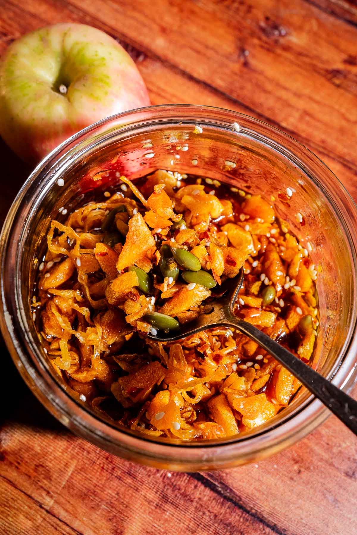 Glass jar filled with shredded apple and seeds mixture on a wooden surface, with a spoon inside, a whole apple next to the jar, and a hint of apple chili crisp for an added kick.