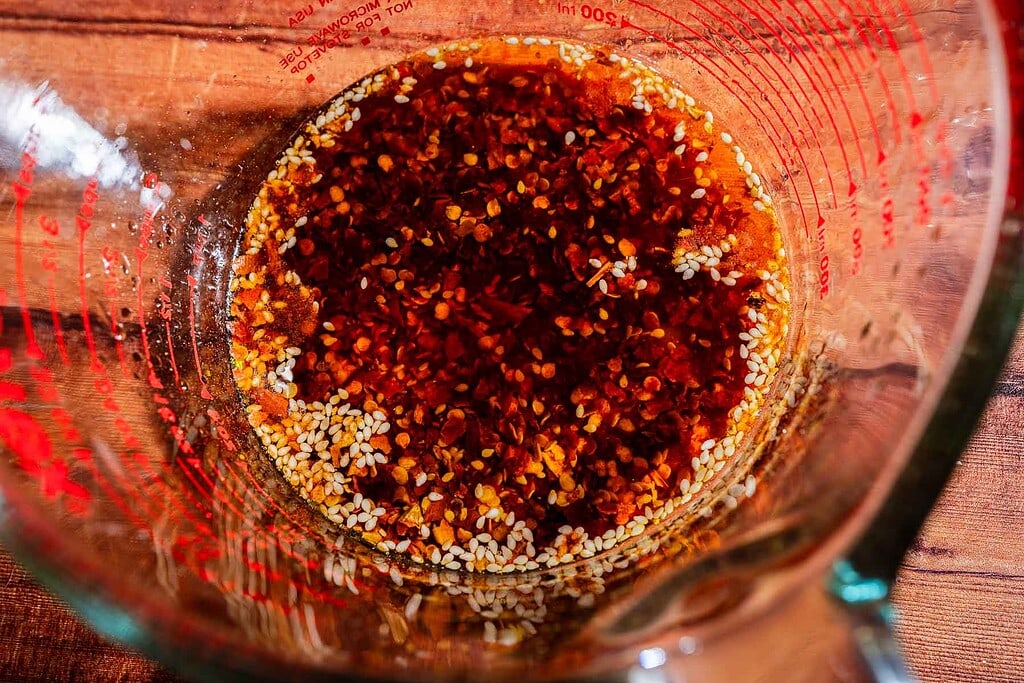 Glass measuring cup containing a mixture of oil, red chili flakes, sesame seeds, and a hint of apple chili crisp on a wooden surface.