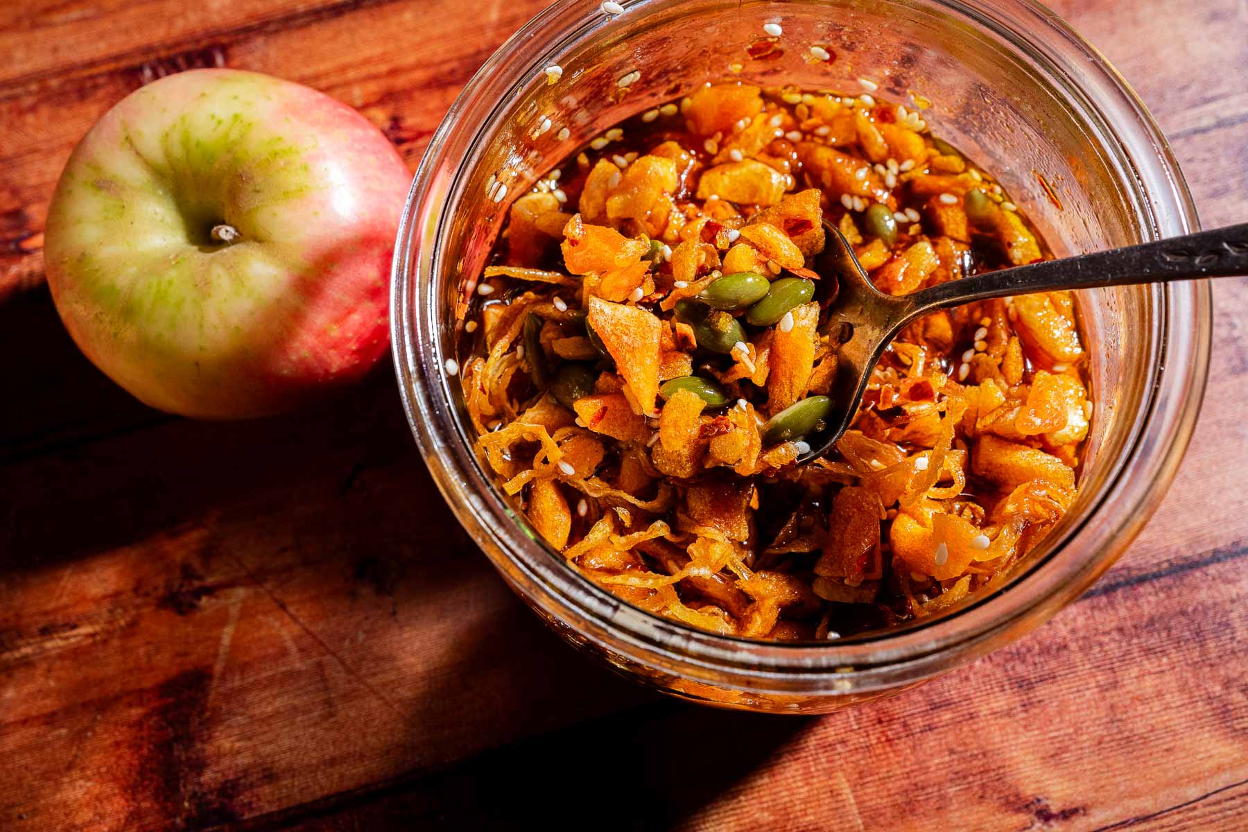 Glass jar of apple chutney with pumpkin seeds and a hint of apple chili crisp, with a spoon next to a whole apple on a wooden surface, lit by sunlight.