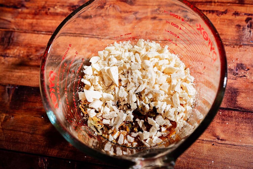 A glass measuring cup filled with light-colored, chopped or crumbled pieces of apple chili crisp sits on a wooden surface.