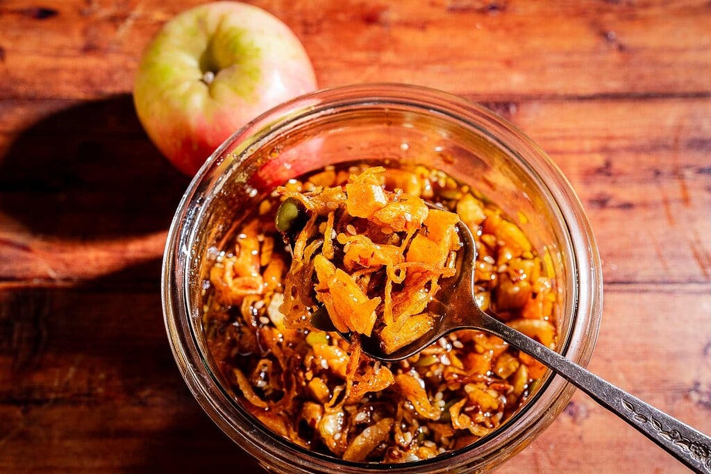 A spoon lifts a portion of chunky apple chili crisp chutney from a glass jar, with a whole apple and wooden table in the background.