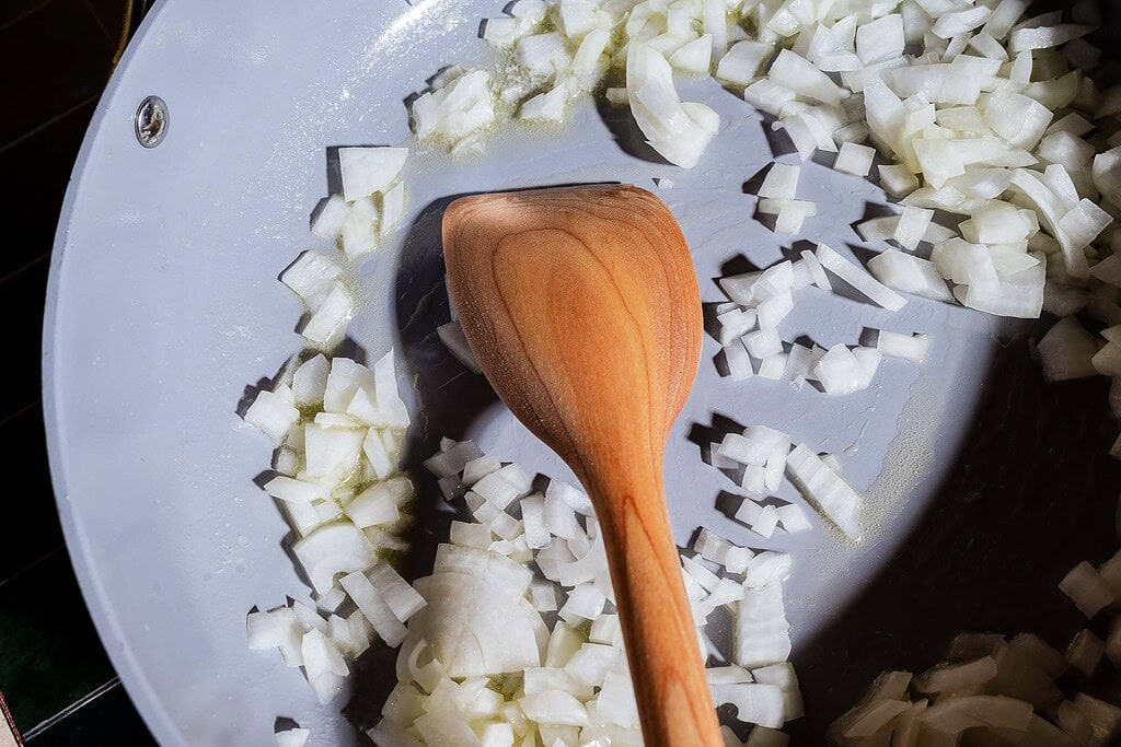 Diced onions are being sautéed in a frying pan with a wooden spoon, ready to be combined with creamy gochujang butter beans for a flavorful twist.