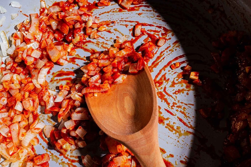 Chopped onions and tomato paste are being stirred in a pan with a wooden spoon, as gochujang butter beans wait to be added for a rich, savory twist.