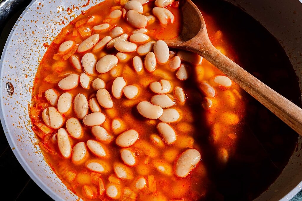 White beans simmer in a vibrant, gochujang-infused tomato-based sauce with chopped onions in a white pan. A wooden spoon rests on the side of the pan, ready to serve these flavorful gochujang butter beans.