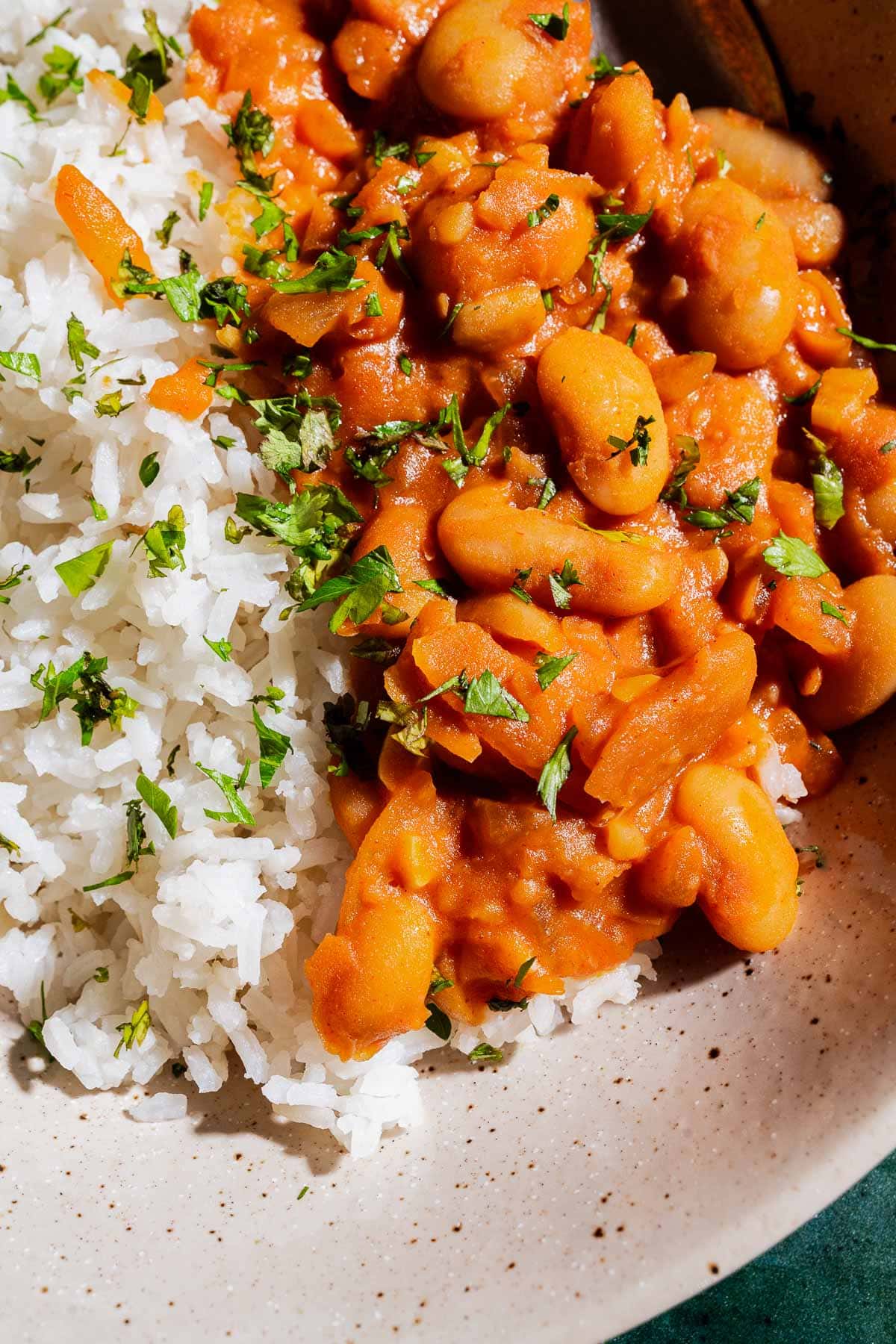 A close-up of white rice and gochujang butter beans stewed in a tomato-based sauce, garnished with chopped herbs, served on a beige plate.