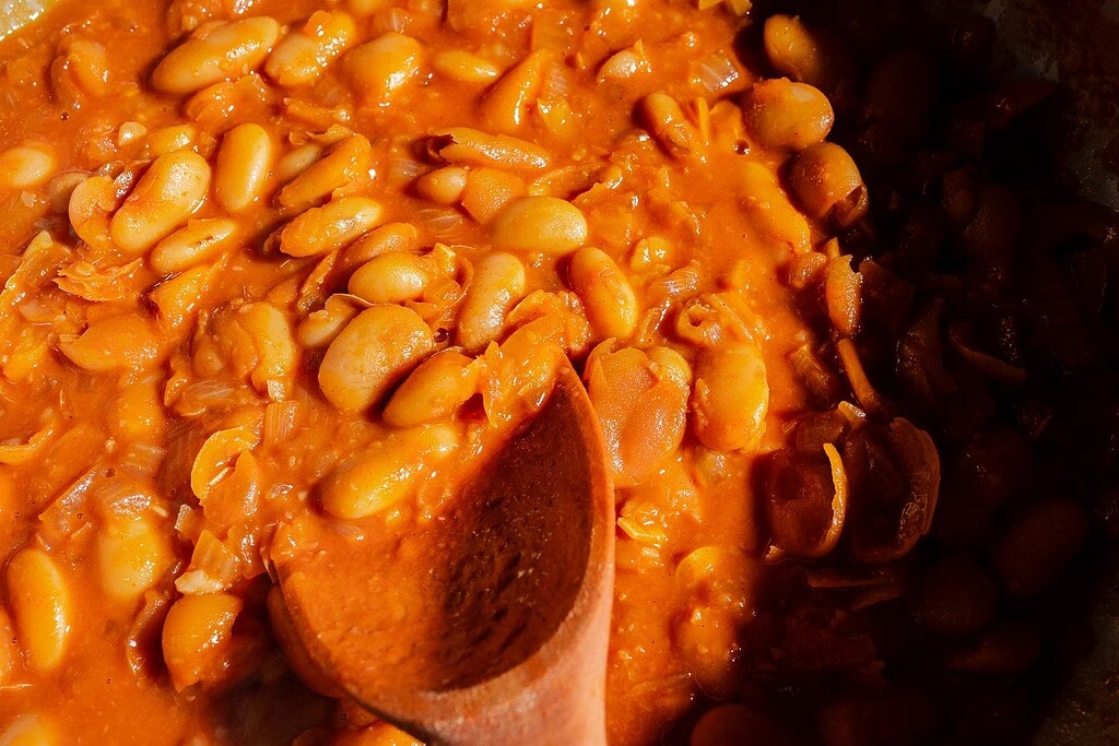 A close-up of cooked gochujang butter beans in a thick orange sauce, being stirred with a wooden spoon.