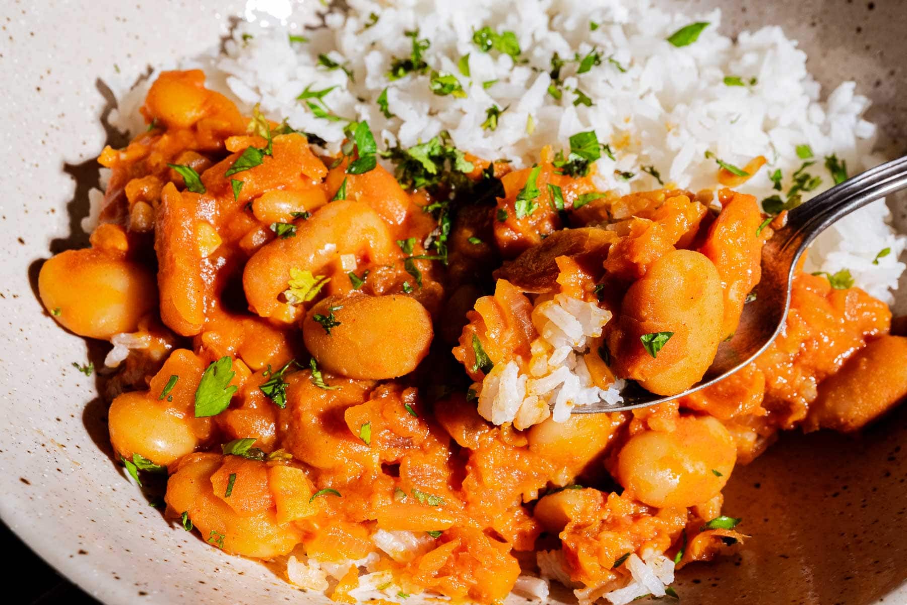 A bowl of white rice and stewed gochujang butter beans in a tomato-based sauce, garnished with chopped herbs, with a spoon scooping a portion.