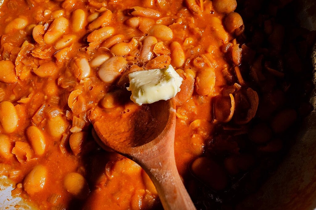 A wooden spoon holds a pat of gochujang butter over a pot of baked beans in a thick tomato sauce.