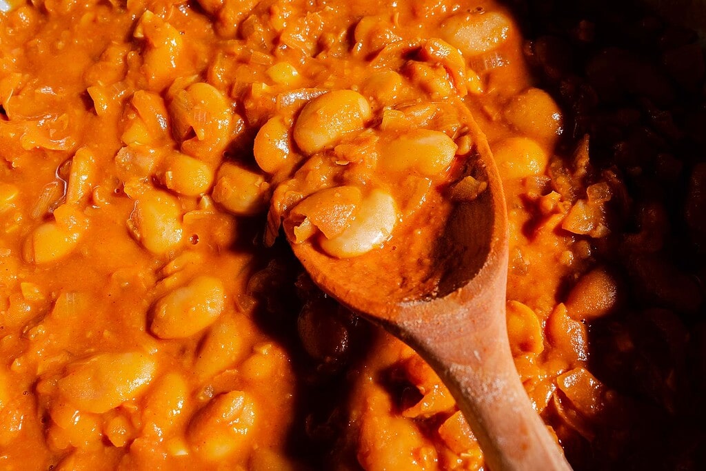 Close-up of a wooden spoon stirring creamy gochujang butter beans in a thick, orange-colored sauce.