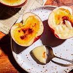 Two halved apples filled with creamy custard and caramelized tops—a delightful twist on apple creme brulee—sit on a speckled plate; a spoon with custard rests beside them. A newspaper is partially visible in the background.