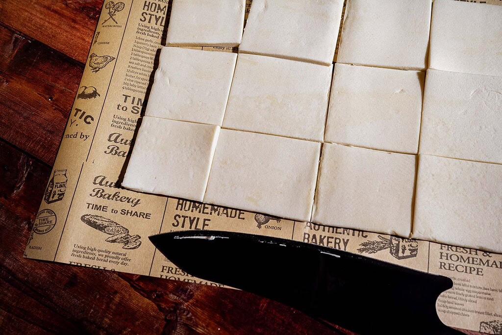 Squares of uncooked hatch chile puff pastry dough are arranged on parchment paper, with a black knife placed partially underneath on a wooden surface.