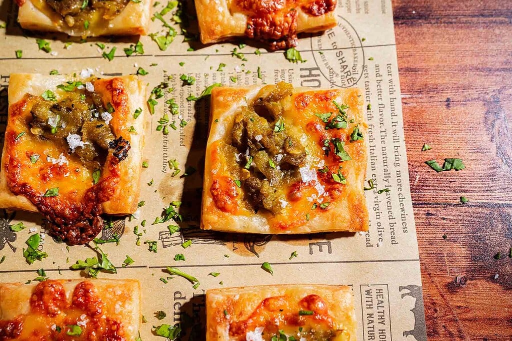 Square hatch chile puff pastry pieces topped with cheese, green chiles, and herbs are arranged on parchment paper with scattered chopped parsley.