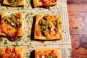 Square hatch chile puff pastry pieces topped with cheese, green chiles, and herbs are arranged on parchment paper with scattered chopped parsley.