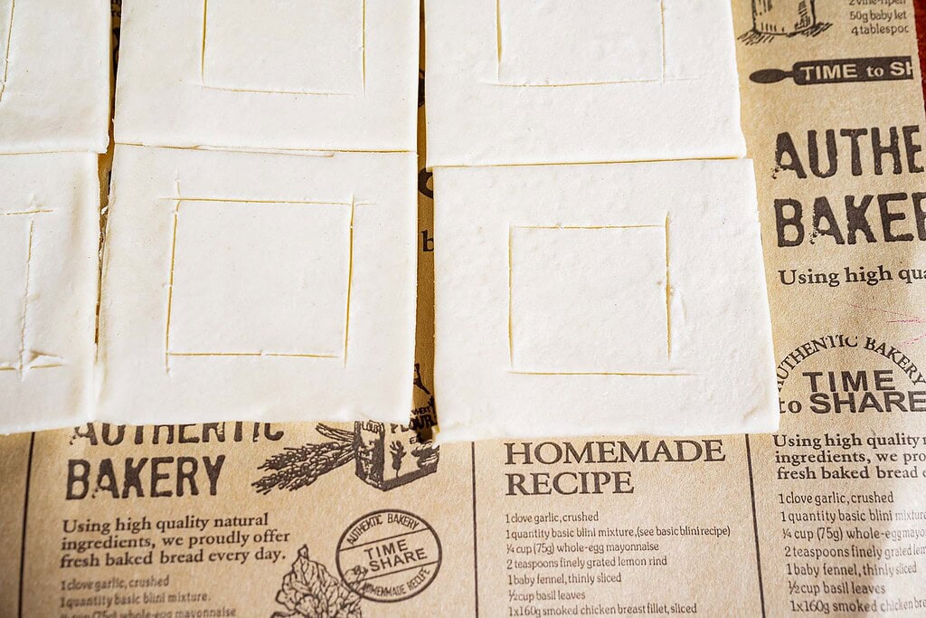 Sheets of raw hatch chile puff pastry with scored square shapes rest on brown paper with bakery-themed text and graphics.