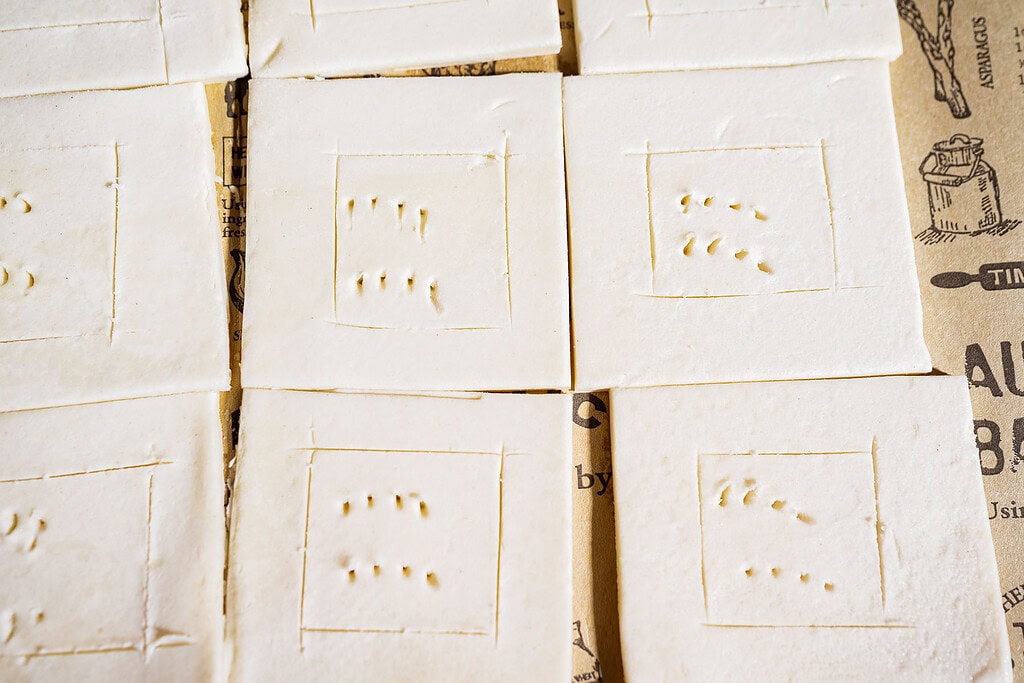 Several square pieces of uncooked hatch chile puff pastry dough with dotted and scored patterns are arranged on printed parchment paper.