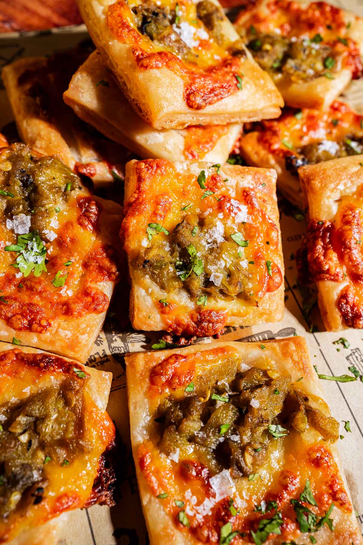 Square hatch chile puff pastry bites topped with melted cheese, diced green chiles, and a sprinkle of herbs and sea salt, arranged on parchment paper.