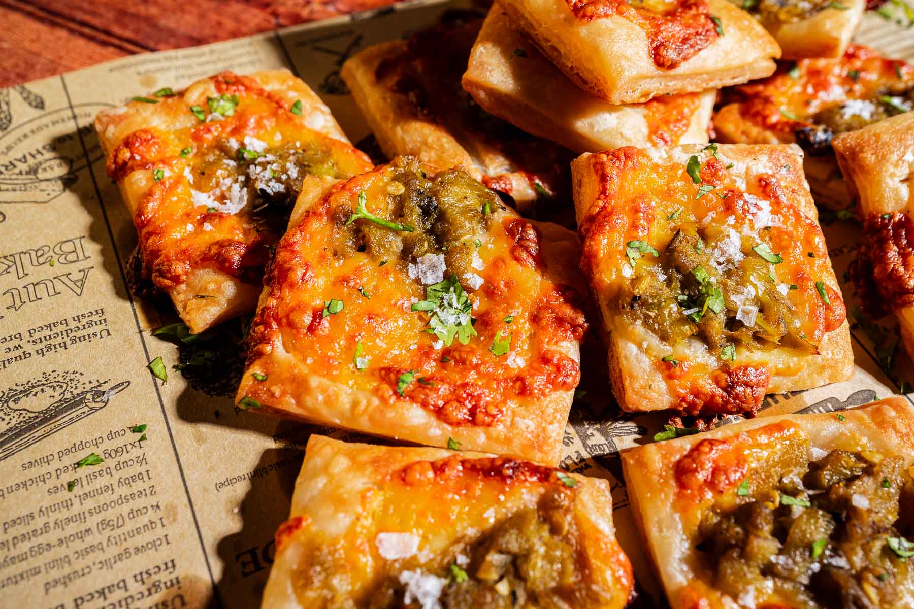 Square puff pastry pieces topped with melted cheese, hatch chiles, herbs, and coarse salt are arranged on newspaper-style parchment paper.