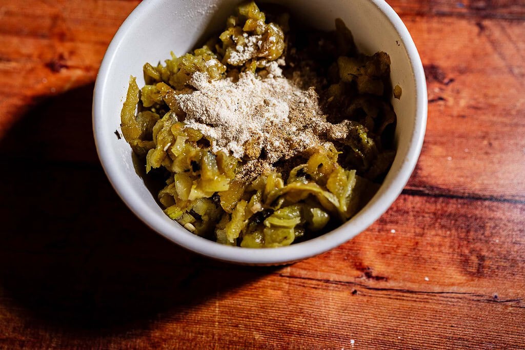 White bowl with chopped green chili peppers and a hint of hatch chile puff pastry aroma, topped with a pile of light brown powdered seasoning, placed on a wooden surface.