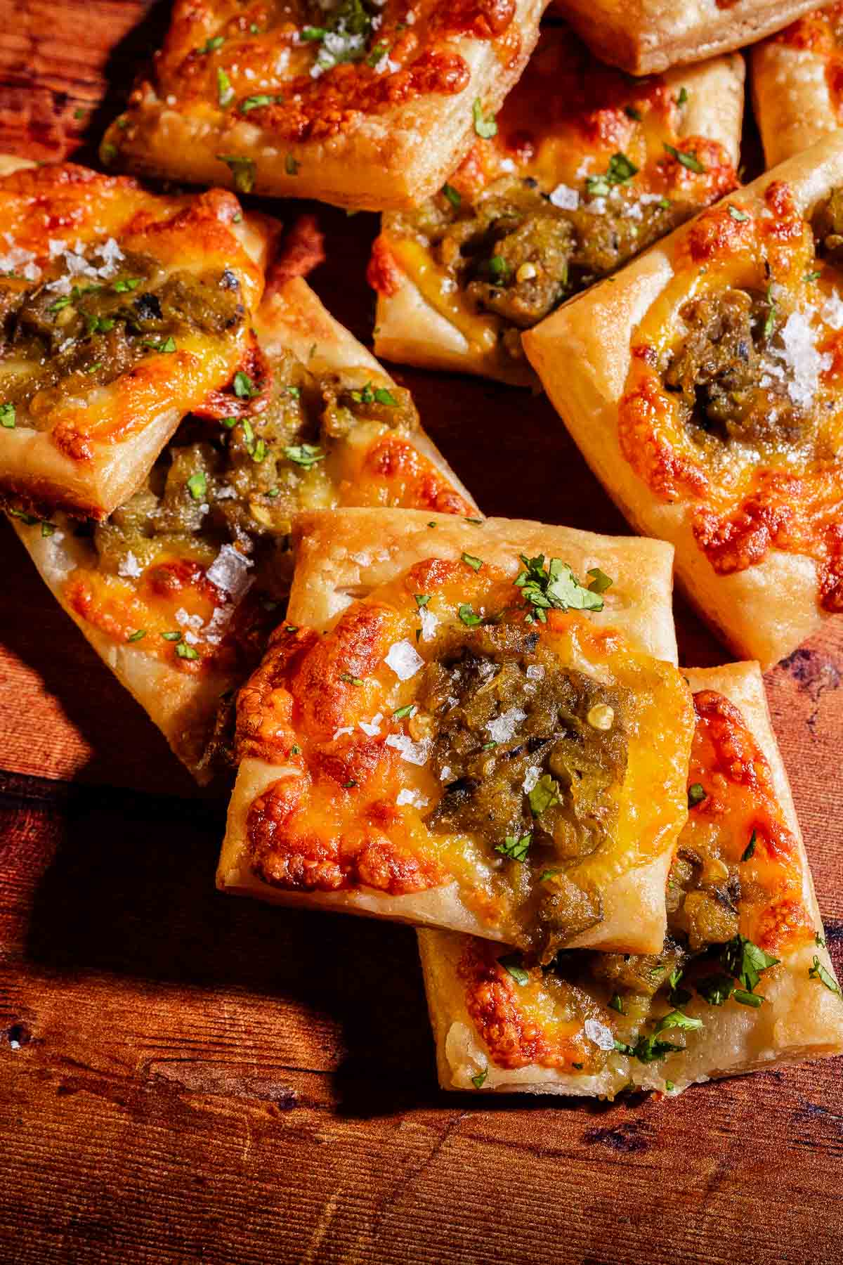 Several rectangular pieces of hatch chile puff pastry, topped with melted cheese, chopped green chiles, and fresh herbs are arranged on a wooden surface.