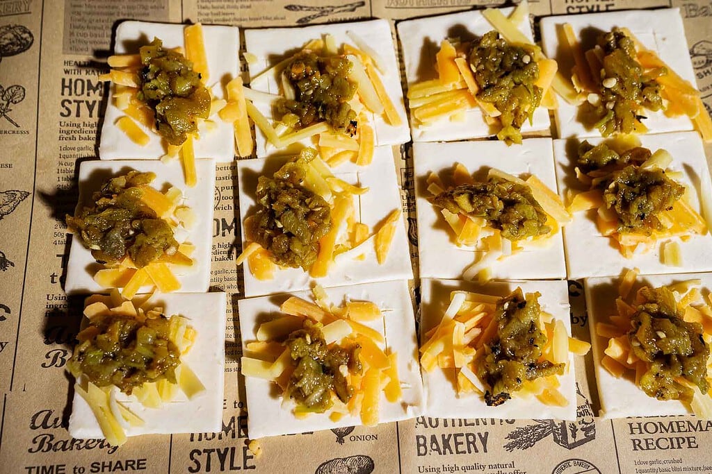 Square crackers topped with shredded yellow and white cheese and a scoop of green relish, arranged in rows on printed parchment paper, bring to mind the bold flavors of hatch chile puff pastry in their vibrant presentation.