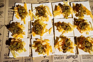 Square crackers topped with shredded yellow and white cheese and a scoop of green relish, arranged in rows on printed parchment paper, bring to mind the bold flavors of hatch chile puff pastry in their vibrant presentation.