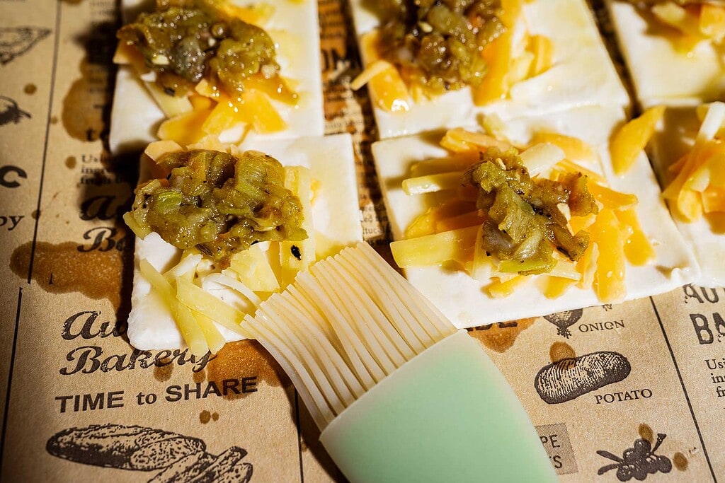 Unbaked crackers topped with shredded cheese and chopped hatch chile are arranged on a paper surface, reminiscent of a hatch chile puff pastry preparation, with a silicone brush nearby.