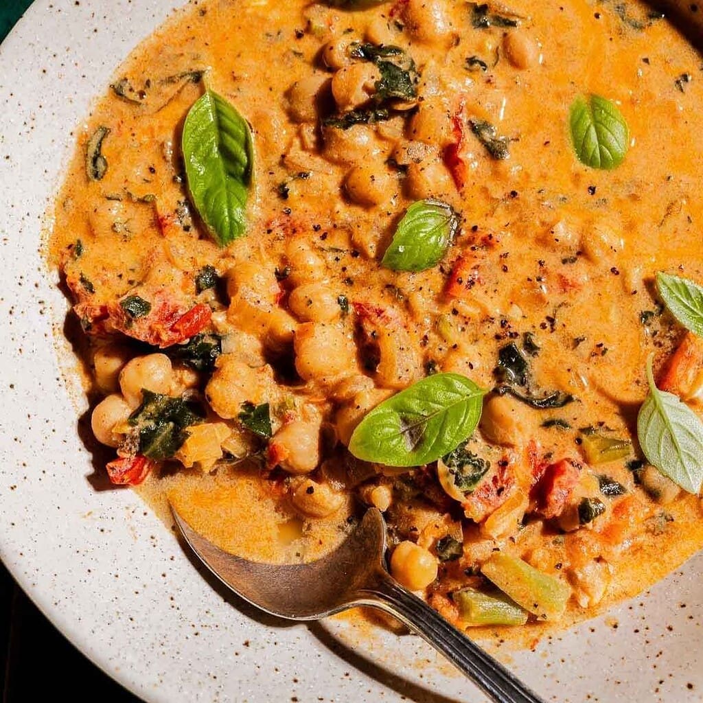 A bowl of creamy chickpea soup with tomatoes, greens, and fresh basil leaves, served with a spoon.
