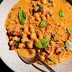 A bowl of creamy chickpea soup with tomatoes, greens, and fresh basil leaves, served with a spoon.