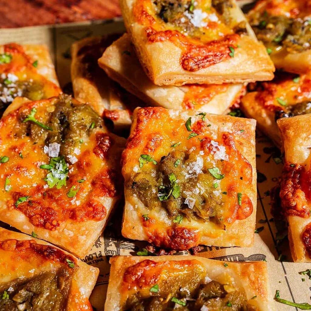Square pieces of baked bread or hatch chile puff pastry topped with melted cheese, green chilies, herbs, and coarse salt, arranged closely on parchment paper.