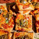 Square pieces of baked bread or hatch chile puff pastry topped with melted cheese, green chilies, herbs, and coarse salt, arranged closely on parchment paper.