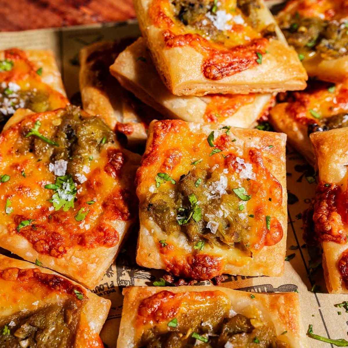 Square pieces of baked bread or hatch chile puff pastry topped with melted cheese, green chilies, herbs, and coarse salt, arranged closely on parchment paper.