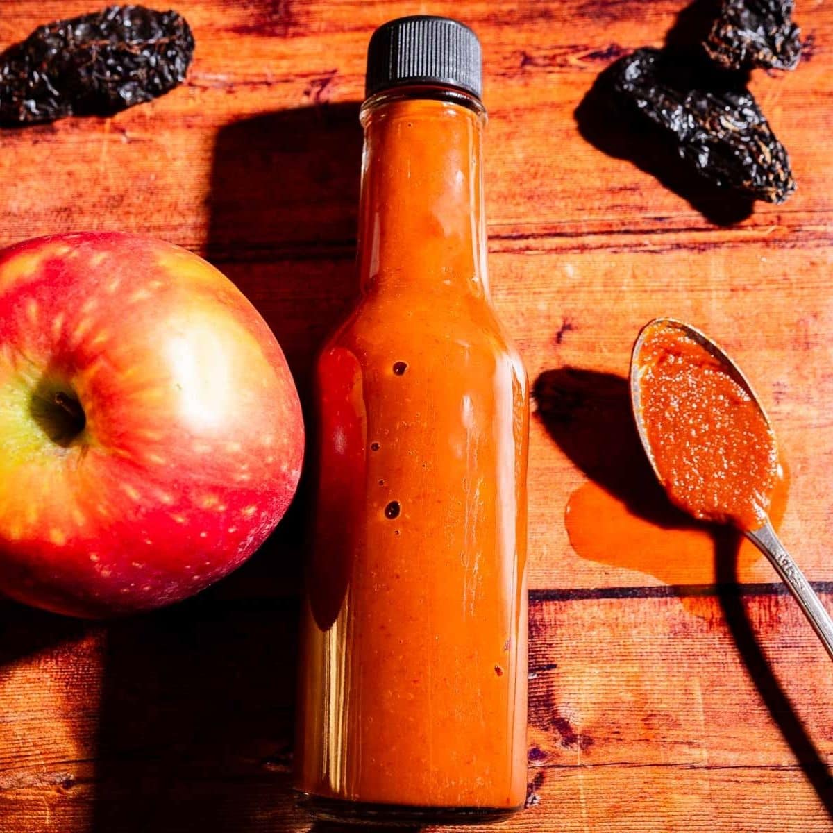 A glass bottle of apple hot sauce sits on a wooden surface beside a red apple, dried peppers, and a spoon with spilled sauce.