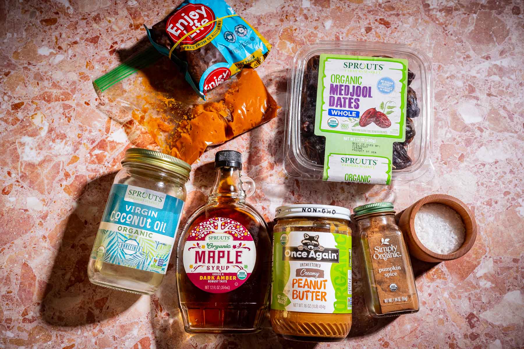 A selection of ingredients on a counter—perfect for making date bark—including coconut oil, maple syrup, peanut butter, Medjool dates, sea salt, a spice mix, and chocolate chips.