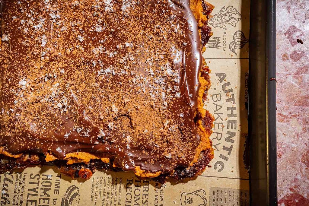 A square dessert bar with a chocolate topping, dusted with cocoa powder and sea salt, sits on branded brown parchment paper in a baking tray—reminiscent of a luxurious date bark treat.