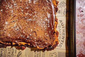 A square dessert bar with a chocolate topping, dusted with cocoa powder and sea salt, sits on branded brown parchment paper in a baking tray—reminiscent of a luxurious date bark treat.