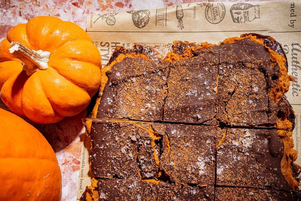 Sliced pumpkin bars with a chocolate topping and a sprinkle of salt, placed on parchment paper next to two small pumpkins—these treats are reminiscent of the popular date bark trend, blending comforting autumn flavors with a satisfying chocolatey finish.