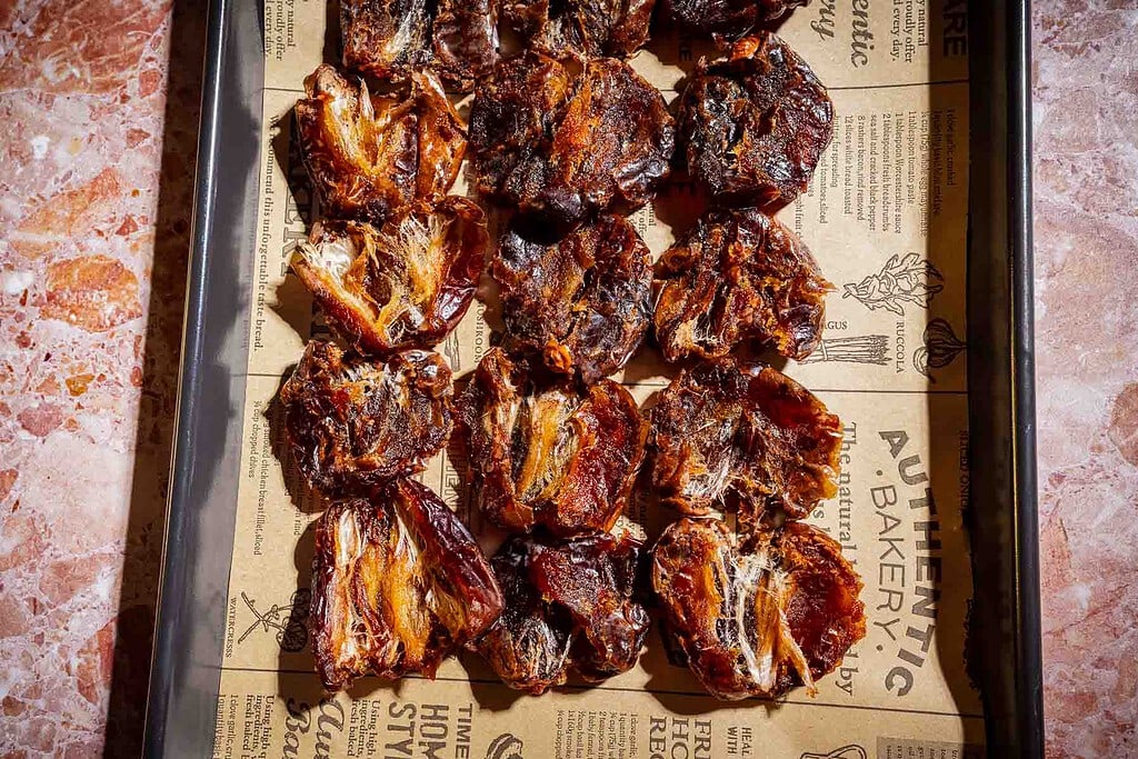A baking tray lined with printed paper holds several pieces of baked or dried fruit, including date bark, arranged in neat rows on a marble surface.