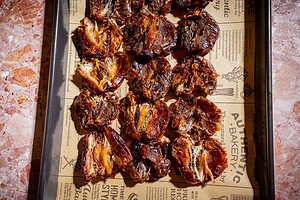 A baking tray lined with printed paper holds several pieces of baked or dried fruit, including date bark, arranged in neat rows on a marble surface.