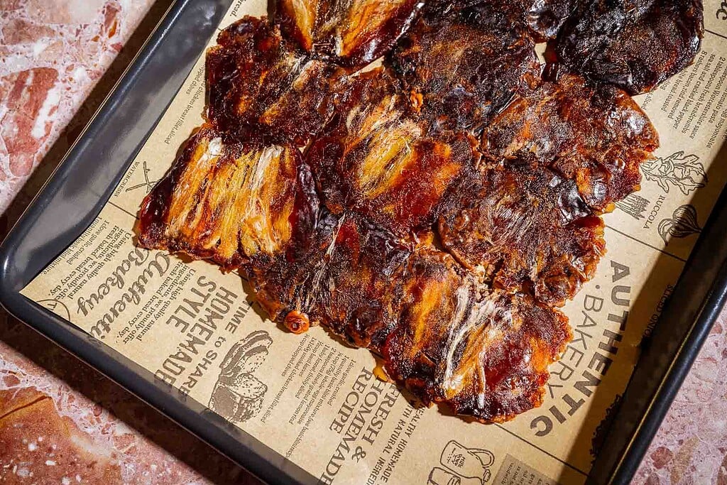Strips of dried, seasoned meat are arranged on a parchment-lined baking tray atop a marble surface, reminiscent of how date bark is carefully prepared and presented for snacking.