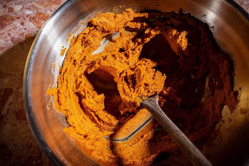 A metal mixing bowl with a wooden spoon containing thick orange-brown dough or batter for date bark, partially mixed, sitting on a reddish marble surface.