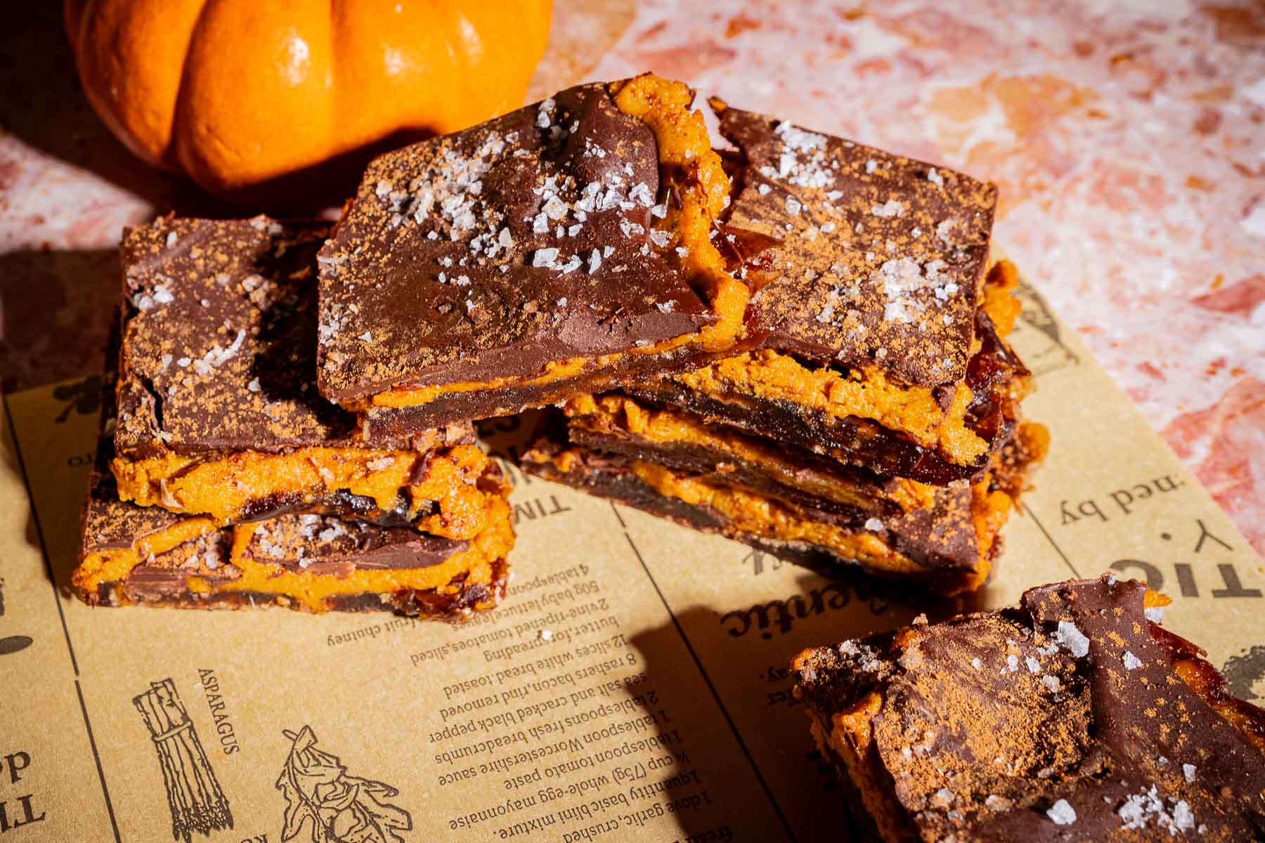 Stacked chocolate and pumpkin bars, reminiscent of date bark, are topped with flaky salt and sit on a sheet of printed paper, with a small decorative pumpkin in the background.
