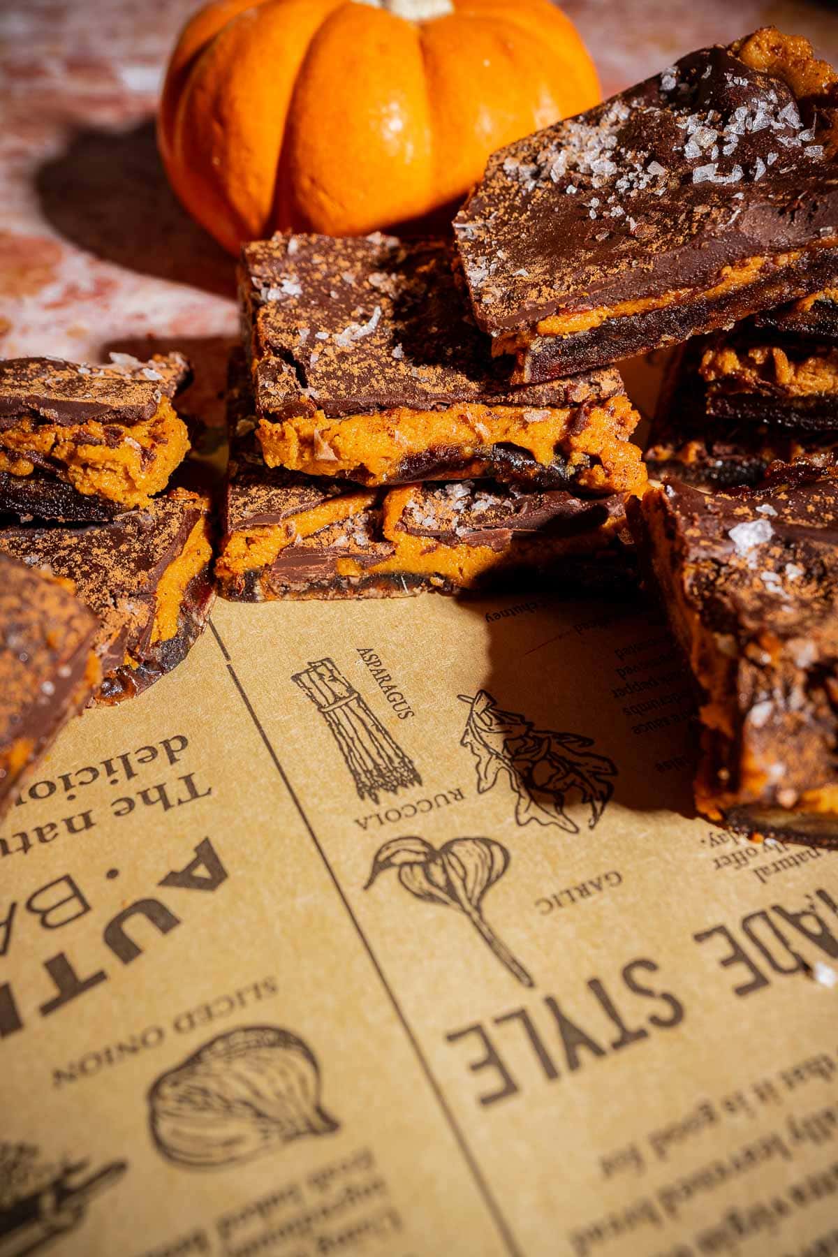 Stacked chocolate and orange pumpkin bars, reminiscent of date bark, are sprinkled with sea salt and displayed on brown paper with food illustrations, accompanied by a small pumpkin in the background.