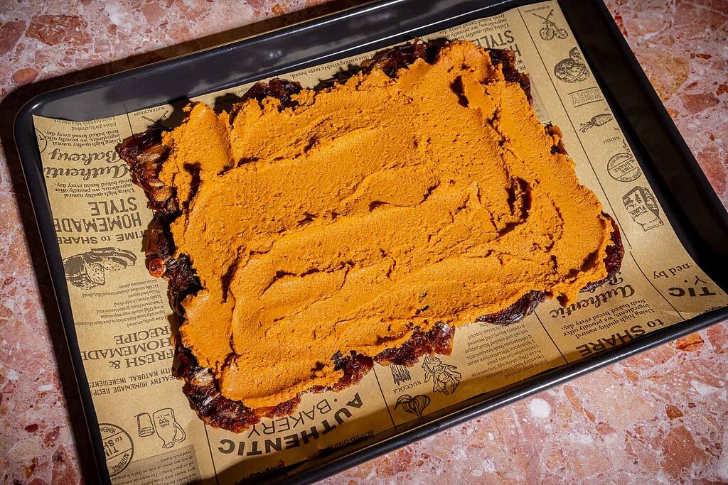 A baking tray lined with printed parchment paper holds a rectangular layer of orange paste spread over a dark, uneven date bark base.