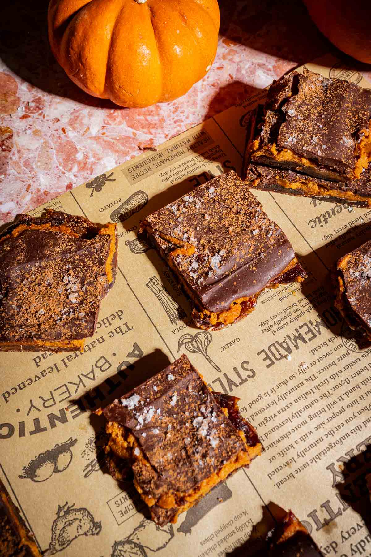 Several chocolate-topped pumpkin bars are arranged on printed parchment paper, reminiscent of classic date bark treats, with a small whole pumpkin in the upper left corner on a marble surface.