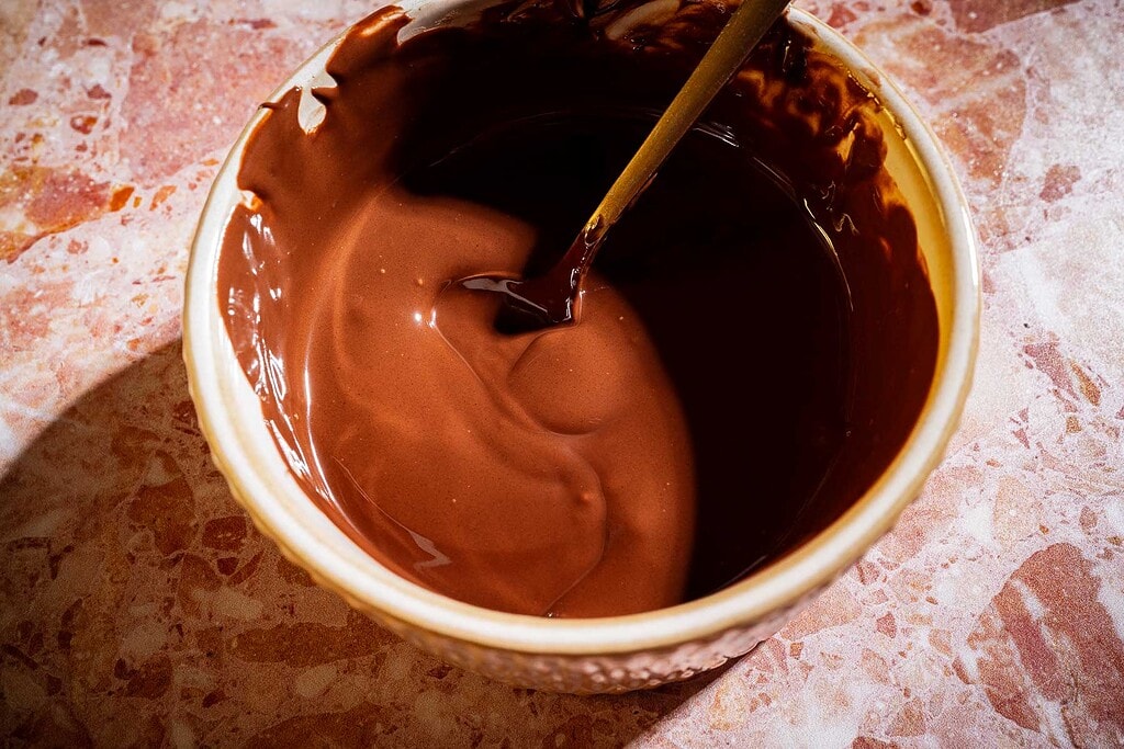A bowl filled with melted chocolate for date bark is being stirred with a wooden spoon, placed on a pink and beige marbled surface.