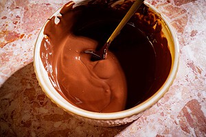A bowl filled with melted chocolate for date bark is being stirred with a wooden spoon, placed on a pink and beige marbled surface.