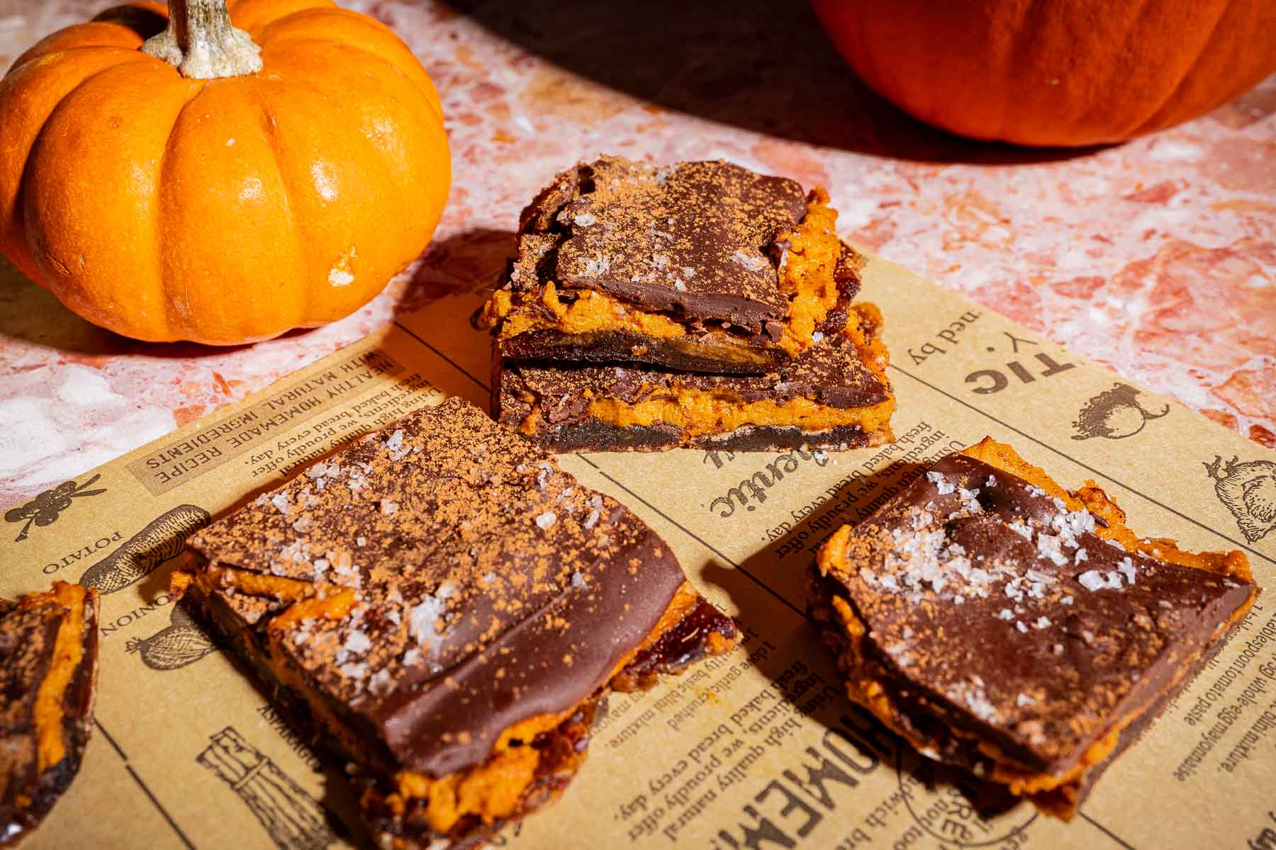 Four chocolate-topped pumpkin bars are arranged on newspaper alongside a small orange pumpkin on a pink marble surface, reminiscent of the trendy date bark treats popular for their rich flavors and eye-catching presentation.