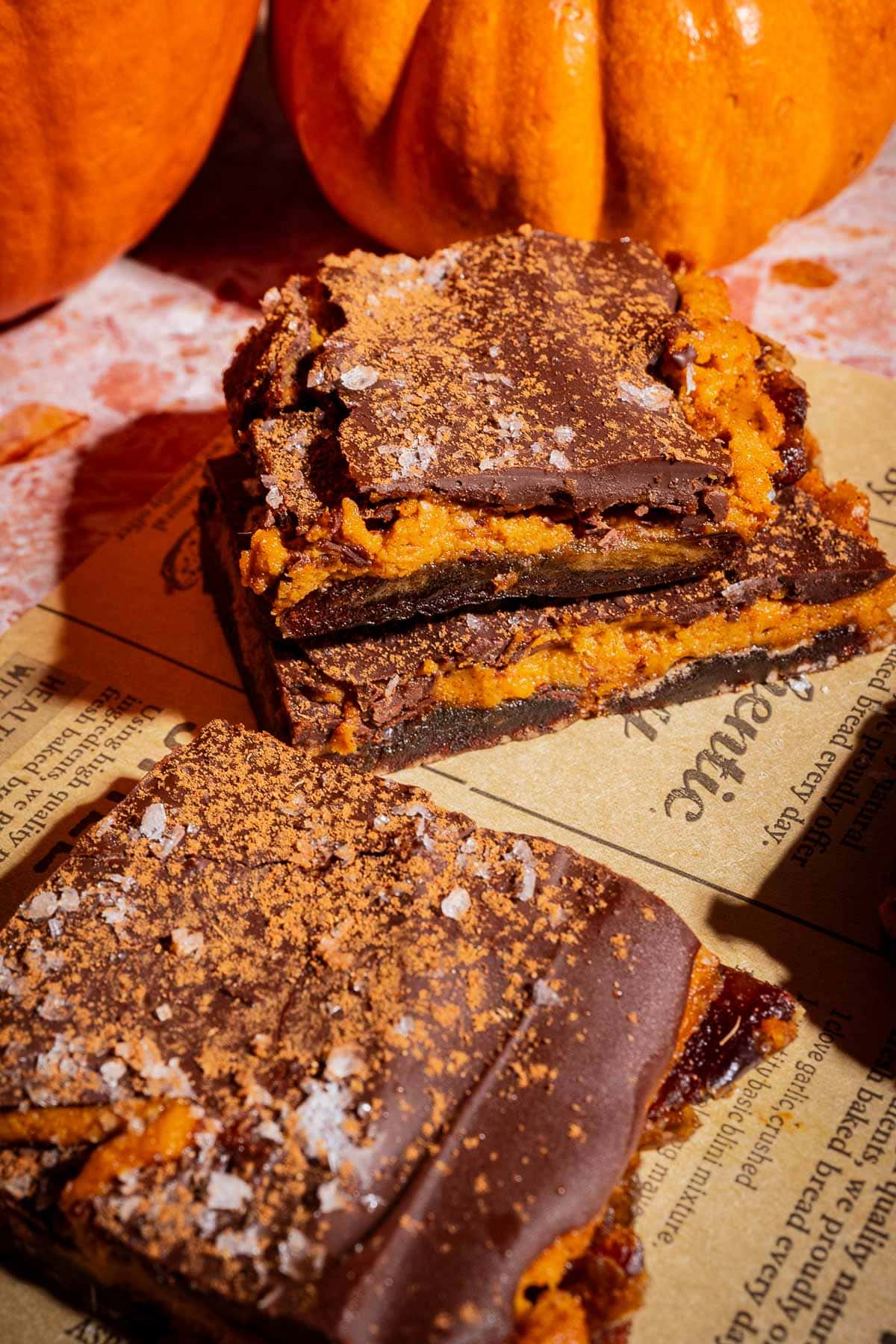 Three chocolate and pumpkin dessert bars, inspired by the popular date bark trend, are stacked on parchment paper with a dusting of cocoa powder and flaky salt, while whole pumpkins sit in the background.